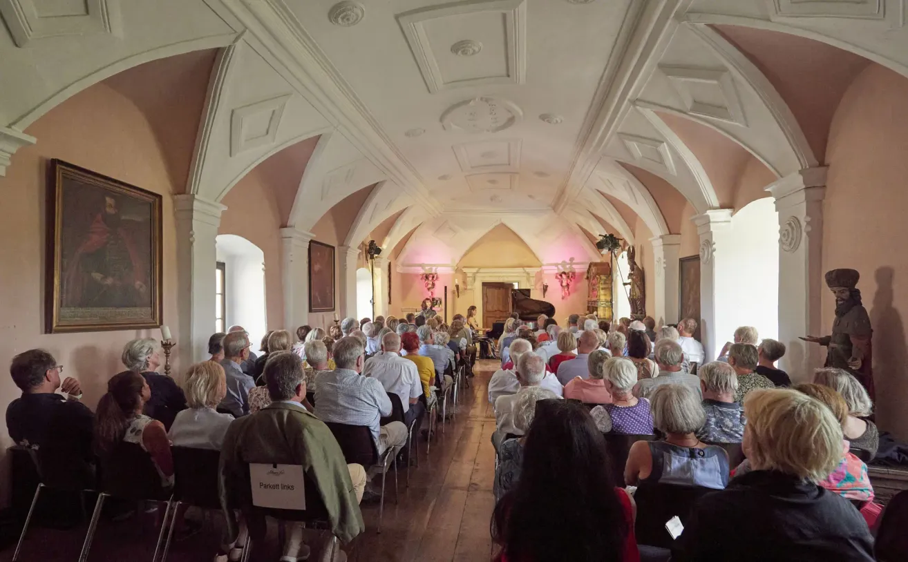 Eine Gruppe von Menschen sitzt in Stühlen in einem Saal und hört eine Klavieraufführung. Der Saal hat gewölbte Decken und Holzböden.