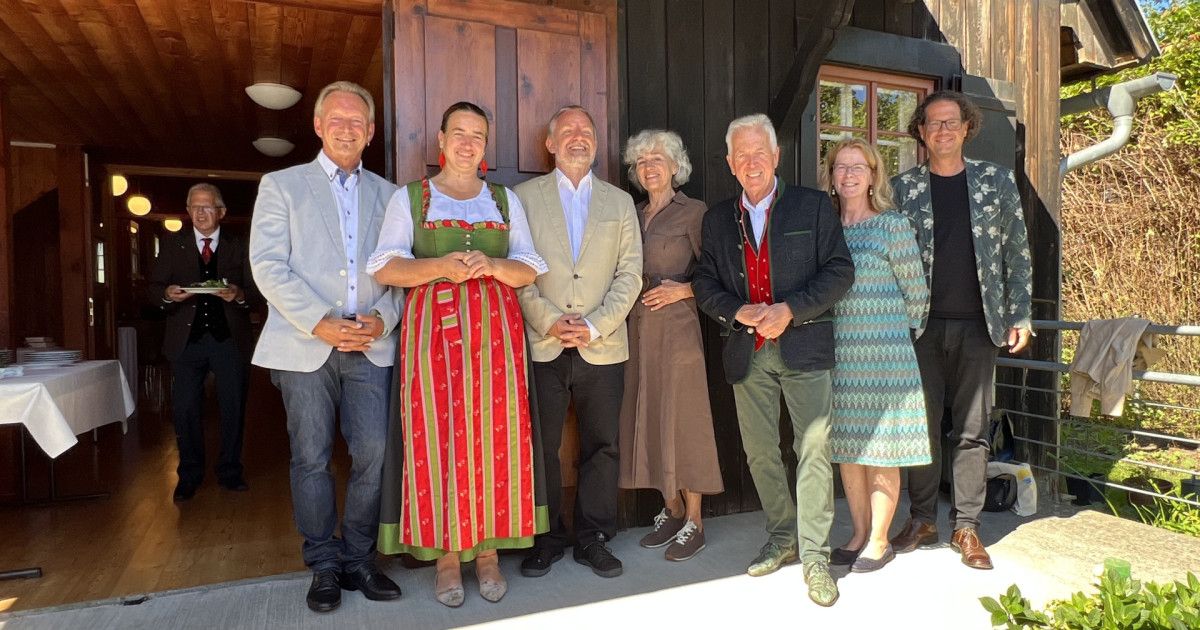 Eine Gruppe von Menschen, in verschiedenen Kleidungsstücken gekleidet, steht vor einem Holzgebäude. Sie lächeln und scheinen für ein Foto zu posieren.