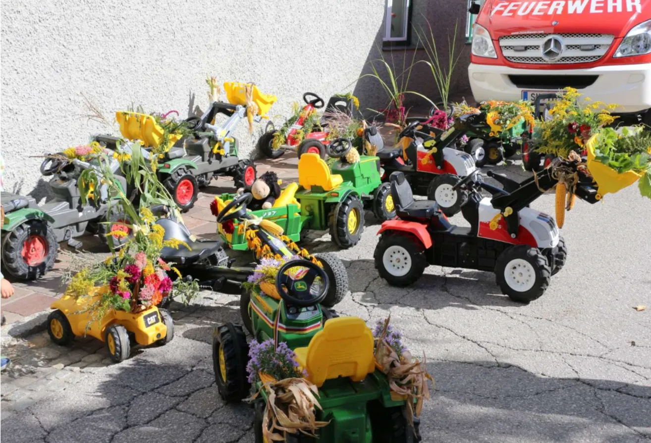 Mehrere Spielzeugtraktoren, jeder mit farbenfrohen Blumen geschmückt, sind in einer Reihe auf einer gepflasterten Oberfläche geparkt. Ein roter Mercedes-Van ist im Hintergrund zu sehen.