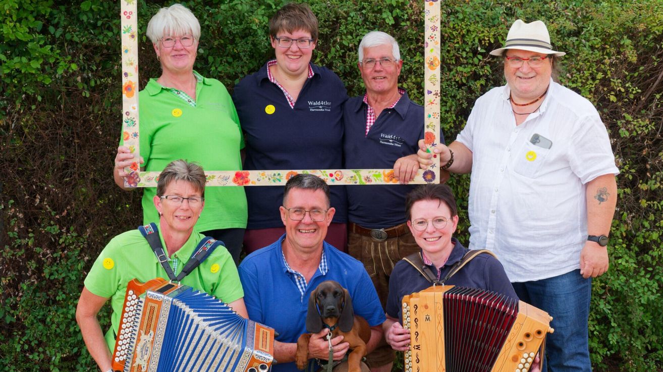 A group of smiling adults, some with musical instruments, pose for a photo in a garden.