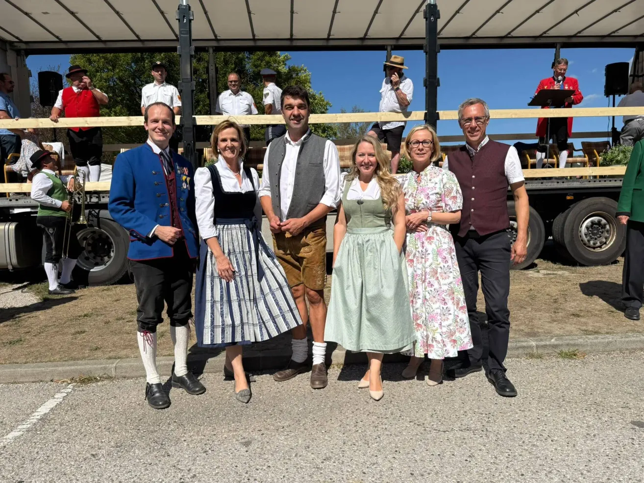 Five individuals dressed in traditional attire stand in front of a wooden structure with a truck in the background.