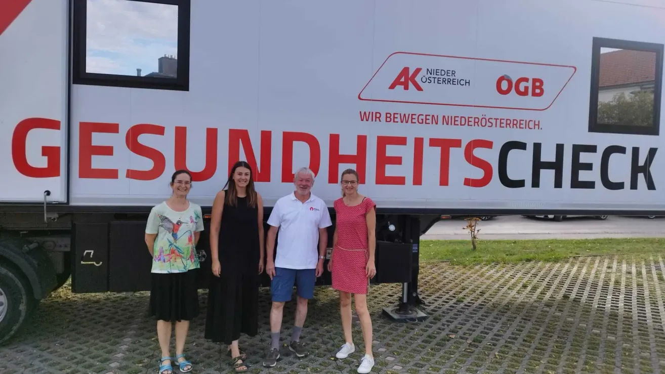 Four individuals stand in front of a white truck with red text reading 'SUNDHEITS'. The man in the middle wears a white shirt and blue shorts, while the women wear black and red dresses. They are all smiling.