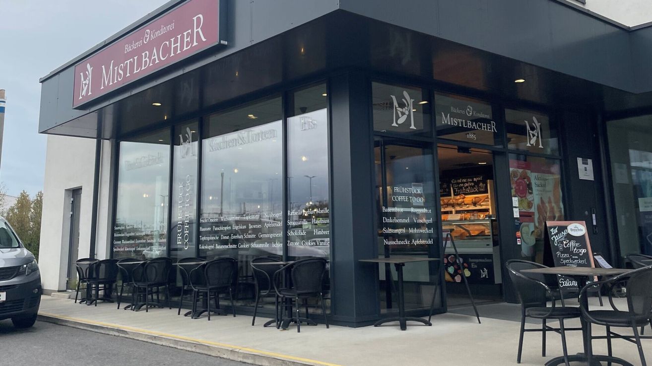 The front of a bakery shop with glass windows and a menu board. Tables and chairs are arranged outside. The shop has a sign that reads 'Mistlbächer'.