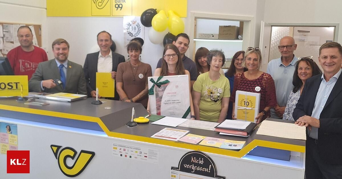 Eine Gruppe von Menschen posiert für ein Foto vor einem Schreibtisch mit Zertifikaten und Papieren. Der Schreibtisch hat ein gelbes und weißes Farbschema. Im Hintergrund befinden sich Luftballons und ein Regal mit Kisten.