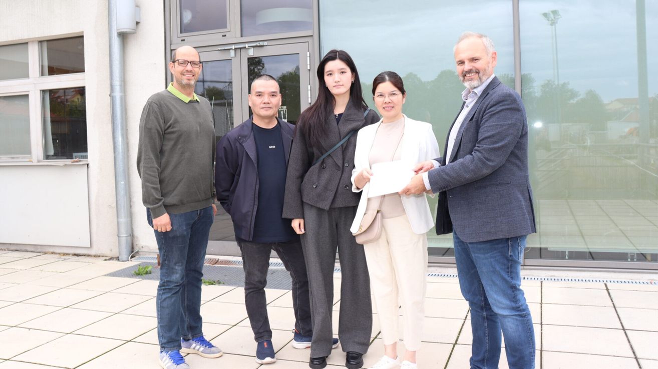 Five people stand in front of a building, smiling. A man on the right hands a paper to a woman in the center.