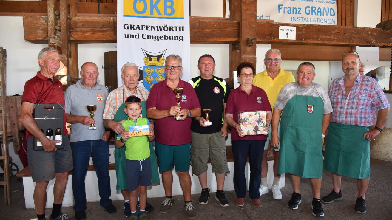 A group of people, including older men and women, are standing together and holding trophies and gifts. They are smiling and posing for a photo. Behind them is a banner with text and a logo.