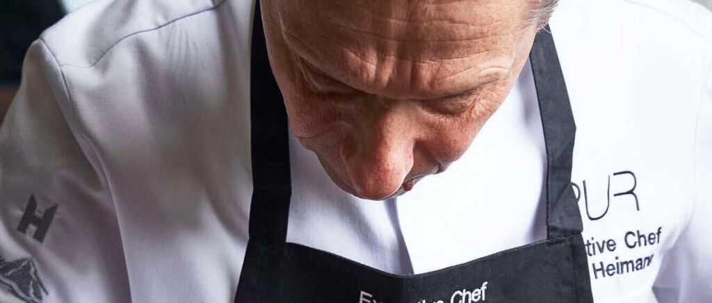 A chef in a white shirt and black apron is concentrating on a task, possibly tasting or preparing food.