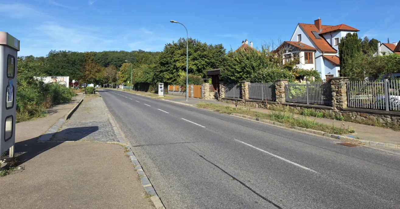 A quiet street with a clear blue sky, lined with trees and houses on either side. The road is empty, and there are streetlights and a small box on the sidewalk.