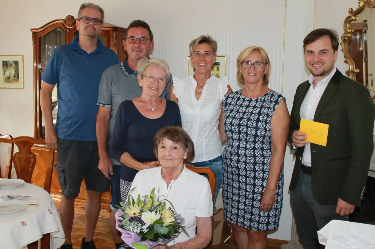 A group of people are gathered for a photo, with a woman in the center holding a bouquet of flowers. Behind her, a woman in a white blouse smiles. On the left, a man in a blue shirt stands next to a woman in glasses. On the right, a man in a suit holds a yellow envelope.