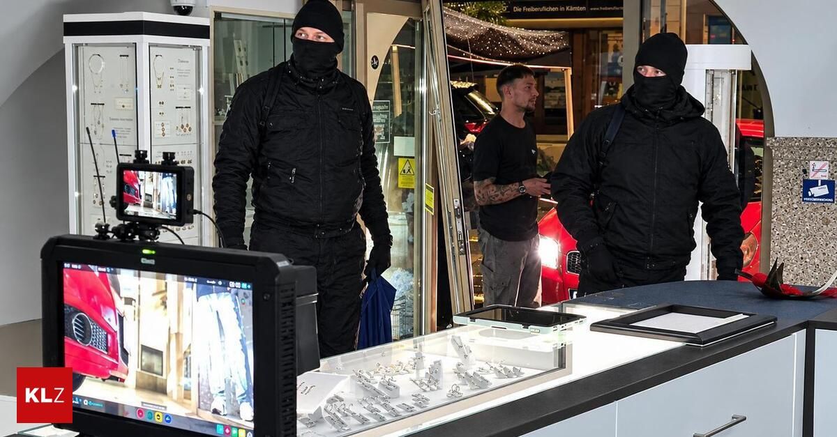 Zwei maskierte Personen in schwarzer Kleidung stehen vor einem Geschäft, während ein Mann in einem schwarzen T-Shirt sie beobachtet. Ein Auto ist draußen geparkt, und ein Monitor zeigt ein Bild an.