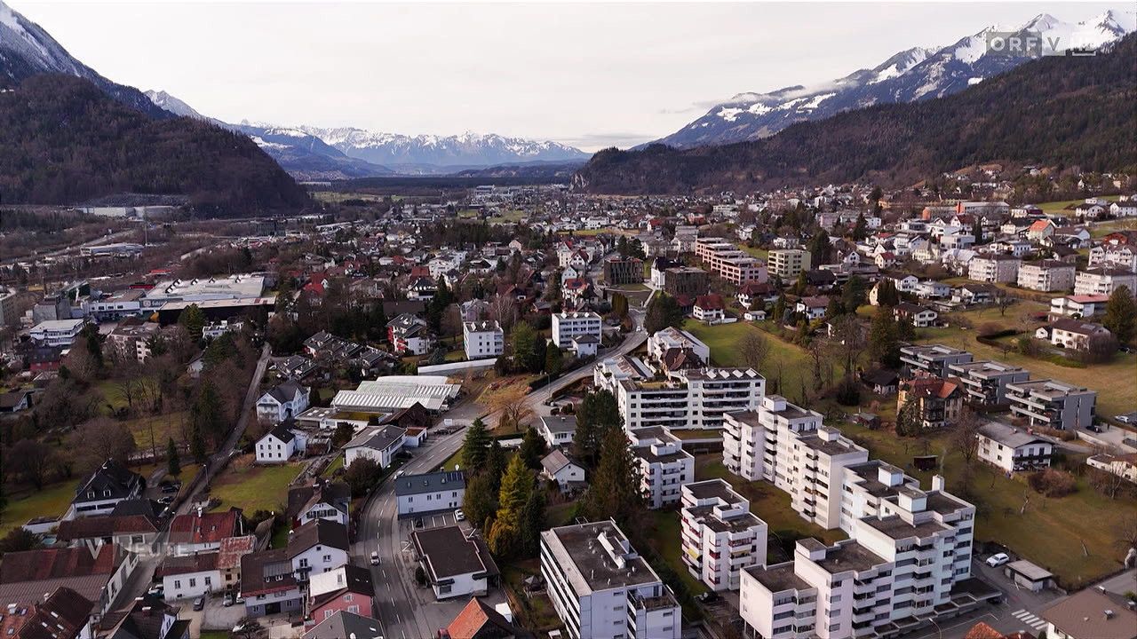 Luftaufnahme einer Stadt mit vielen Gebäuden, umgeben von Bergen und Grünflächen. Die Stadt hat eine Mischung aus Wohn- und Geschäftsgebäuden, einige mit roten Dächern. Die Berge im Hintergrund sind schneebedeckt.