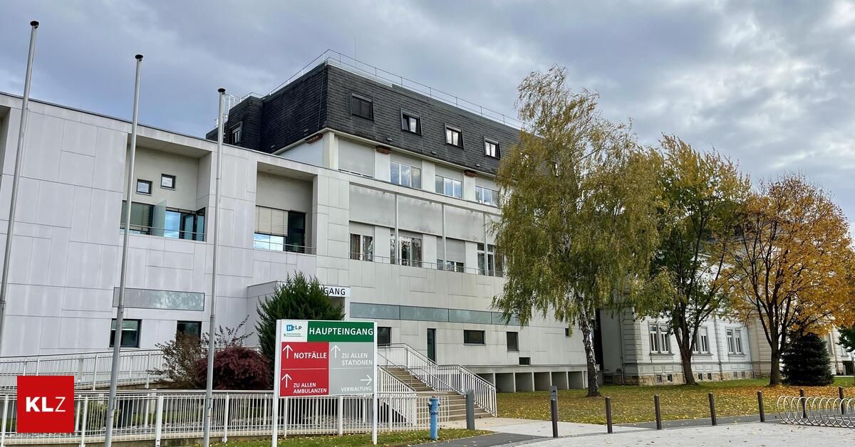 Ein modernes Gebäude mit grauem Dach und weißen Wänden. Es hat viele Fenster und ein Schild, das auf den Haupteingang zeigt. Bäume und Pflanzen stehen vor dem Gebäude.