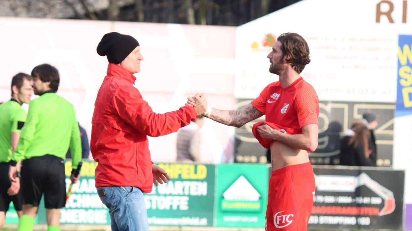 Ein Mann in einem roten Mantel schüttelt einem unbekleideten Fußballspieler in roten Shorts die Hand. Sie stehen vor einem unscharfen Hintergrund mit einem grünen Schild.