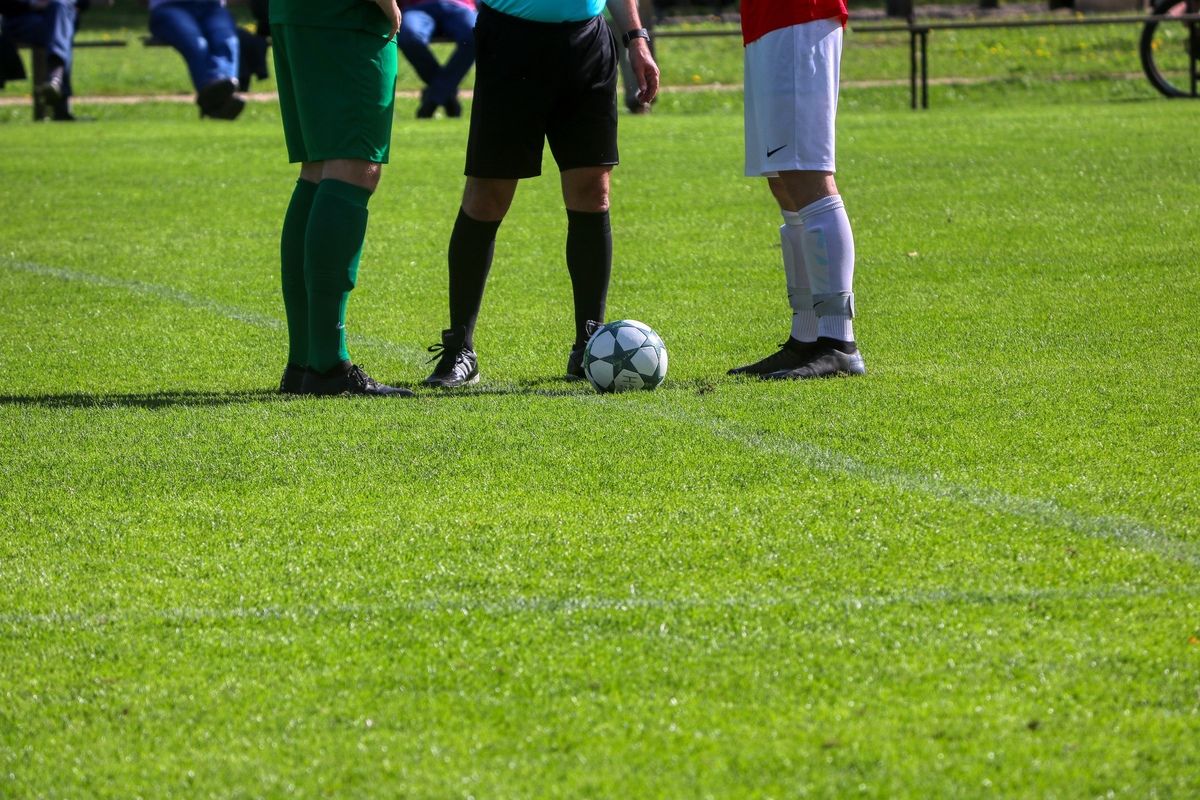 Three soccer players stand on a field with a soccer ball in the center. The player in green is looking at the ball.