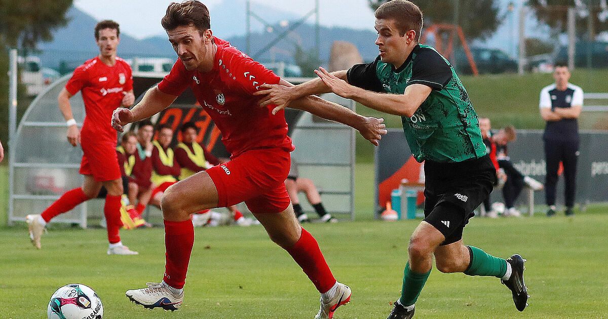 Two soccer players are competing on a field. The player in red is running while the player in green is trying to block him.