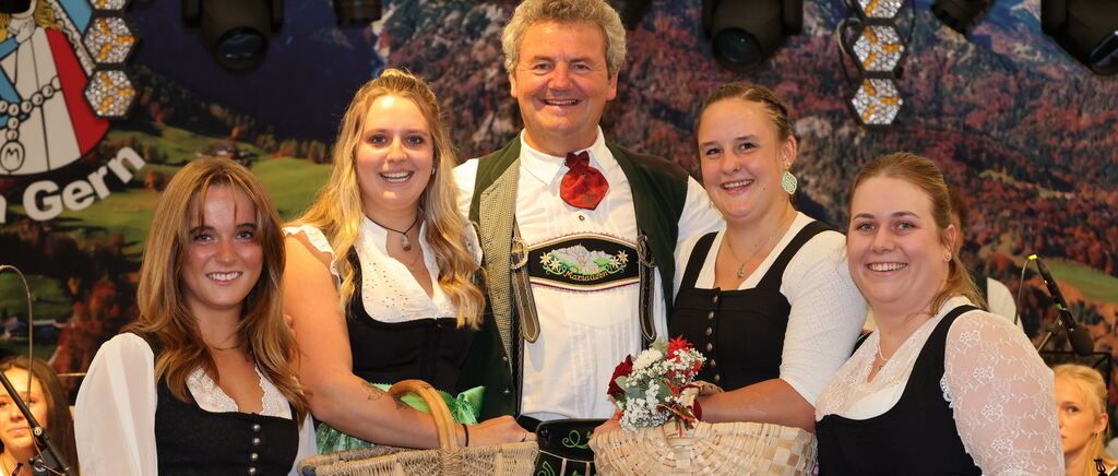 A group of people dressed in traditional German attire, smiling and posing for a photo. The man in the center wears a red bow tie and a green vest. The women on either side of him hold baskets.