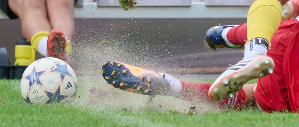 A soccer player kicks a ball on a grassy field, with mud splashing around. The player's cleats are visible, and the ball is in motion.
