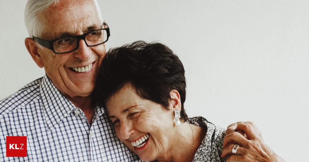 An elderly couple smiling and embracing each other, the man wearing glasses and a checkered shirt, the woman wearing earrings and a necklace.