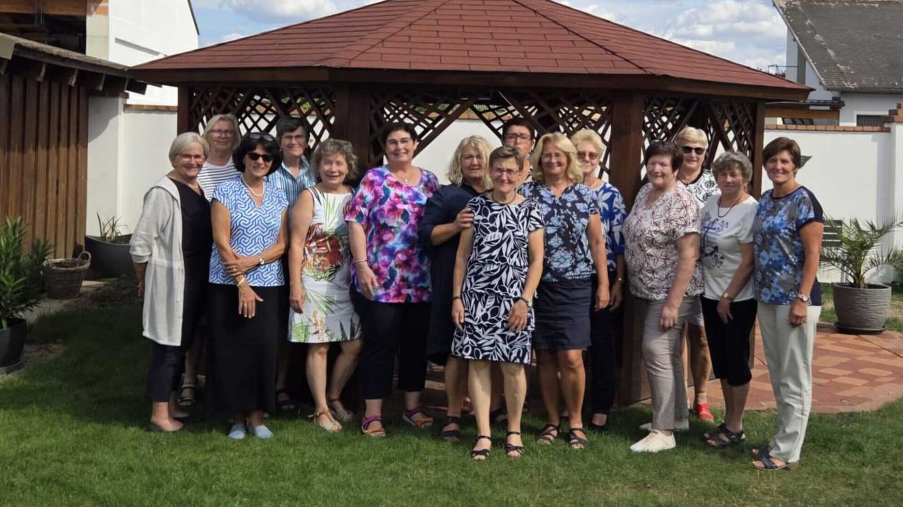 A group of women stands in front of a gazebo, smiling and posing for a photo. They are dressed in various summer outfits.