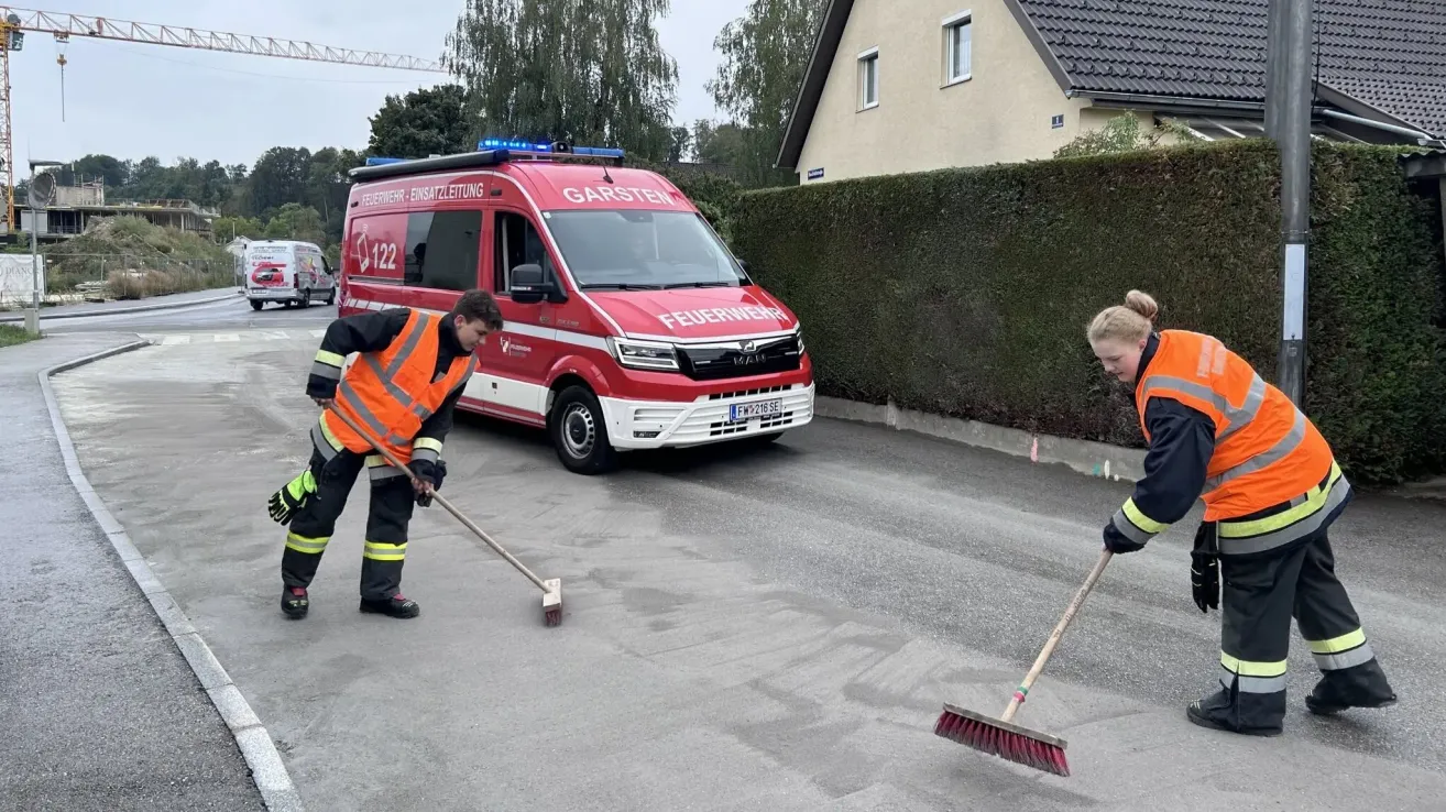 Zwei Feuerwehrleute in orangefarbenen Westen kehren die Straße vor einem roten Feuerwehrauto.