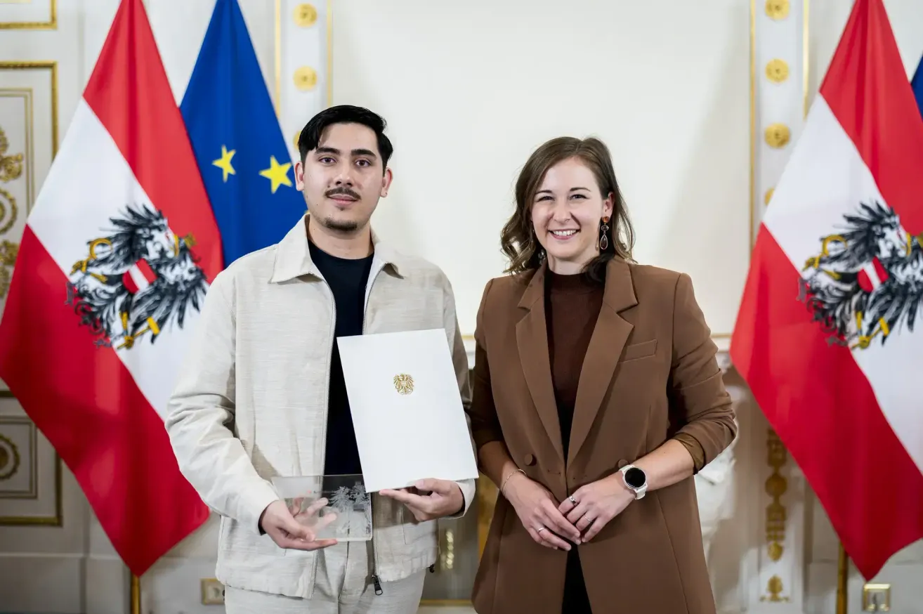 Ein Mann und eine Frau stehen vor Flaggen, lächeln und halten ein Zertifikat. Der Mann trägt eine beige Jacke und die Frau einen braunen Mantel. Hinter ihnen stehen drei Flaggen, darunter die österreichische Flagge.