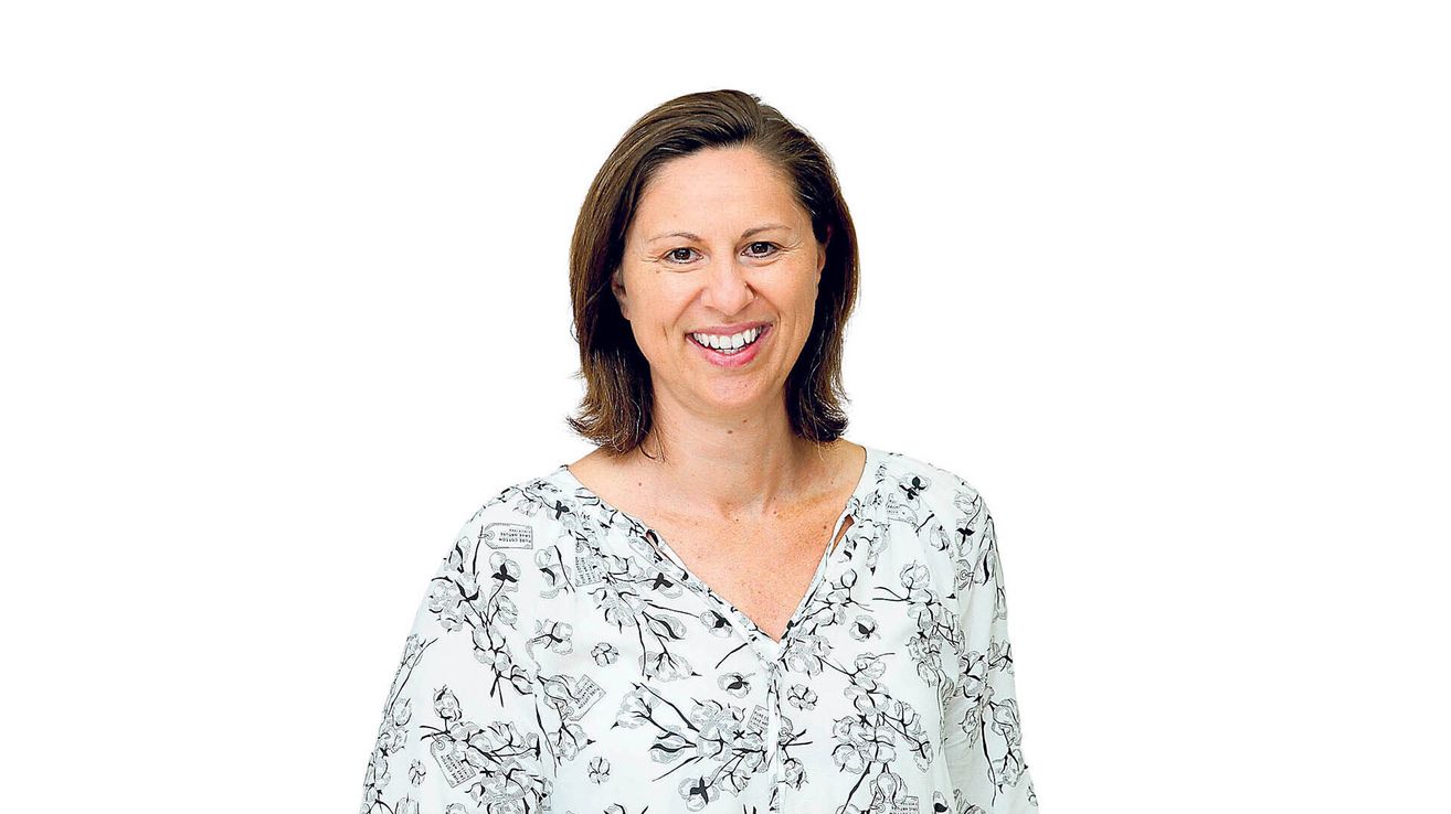 A woman with short hair and a floral shirt smiles at the camera. She is standing against a white background.