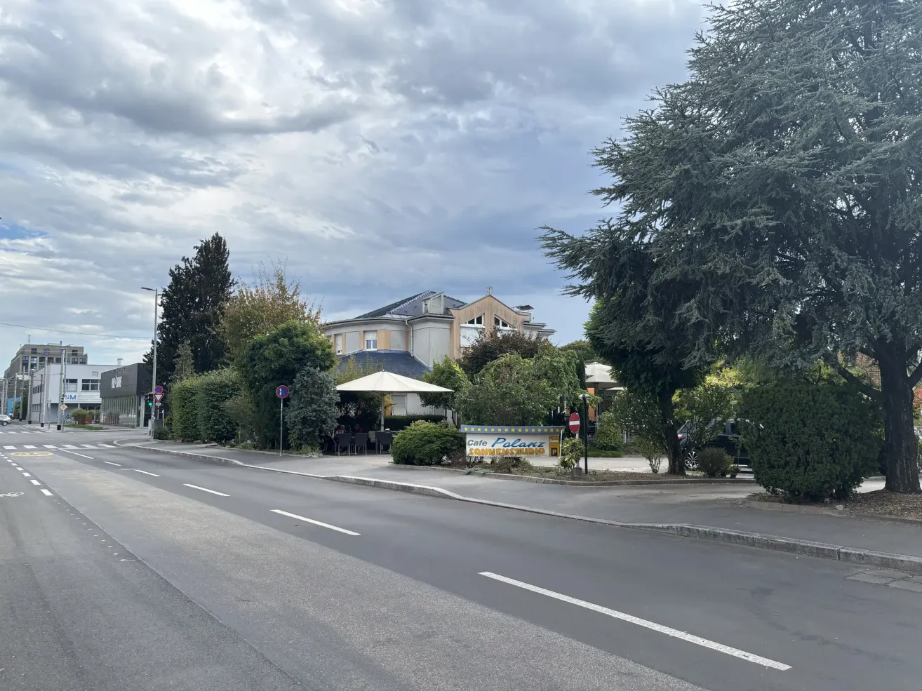 Eine leere Straße mit einem Café-Schild an der Seite, umgeben von Bäumen und Büschen. Im Hintergrund ist ein Gebäude mit einem Giebeldach zu sehen.