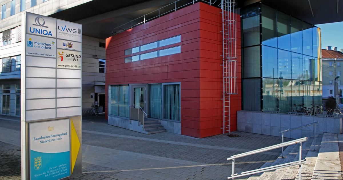 A modern red building with glass windows and a metal ladder on the side. The building has a signboard with the text 'GESUND FIT' on it. There is a staircase leading to the entrance.