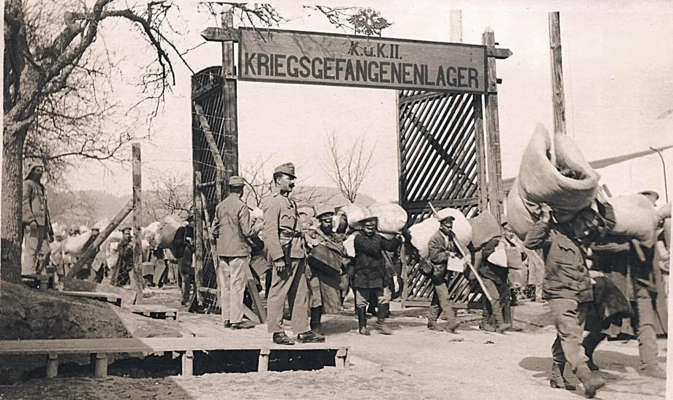 Ein Schwarz-Weiß-Foto von Soldaten, die schwere Lasten durch ein Tor tragen, das Kriegsgefangenenlager beschriftet ist.