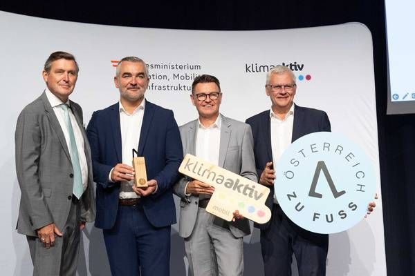 Four men in suits stand on stage, holding awards and signs. One man holds a sign that reads 'klimaaktiv'. Another holds a sign that reads 'Österreich zu Fuß'. The man on the right holds a round sign with the number 4 on it.