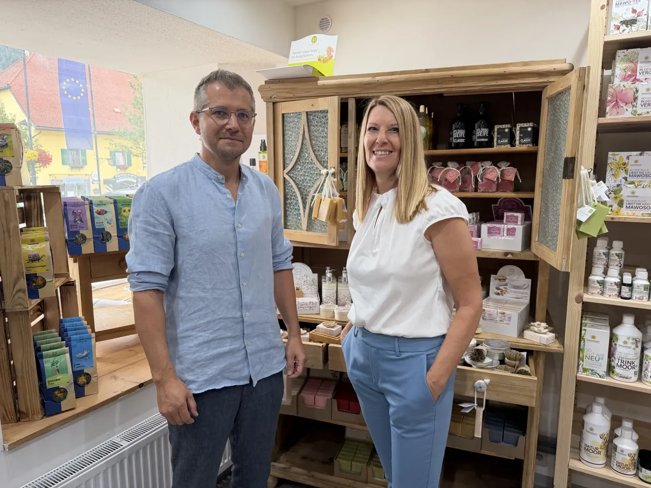 A man and a woman stand in front of a wooden cabinet filled with various products. The man wears a light blue shirt and the woman a white blouse. They both smile for the camera.