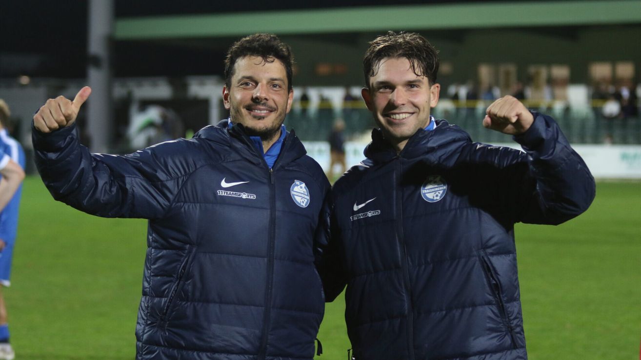Zwei Männer in blauen Jacken mit Nike-Logos und einem Fußballvereinsemblem lächeln und posieren für ein Foto auf einem Feld.