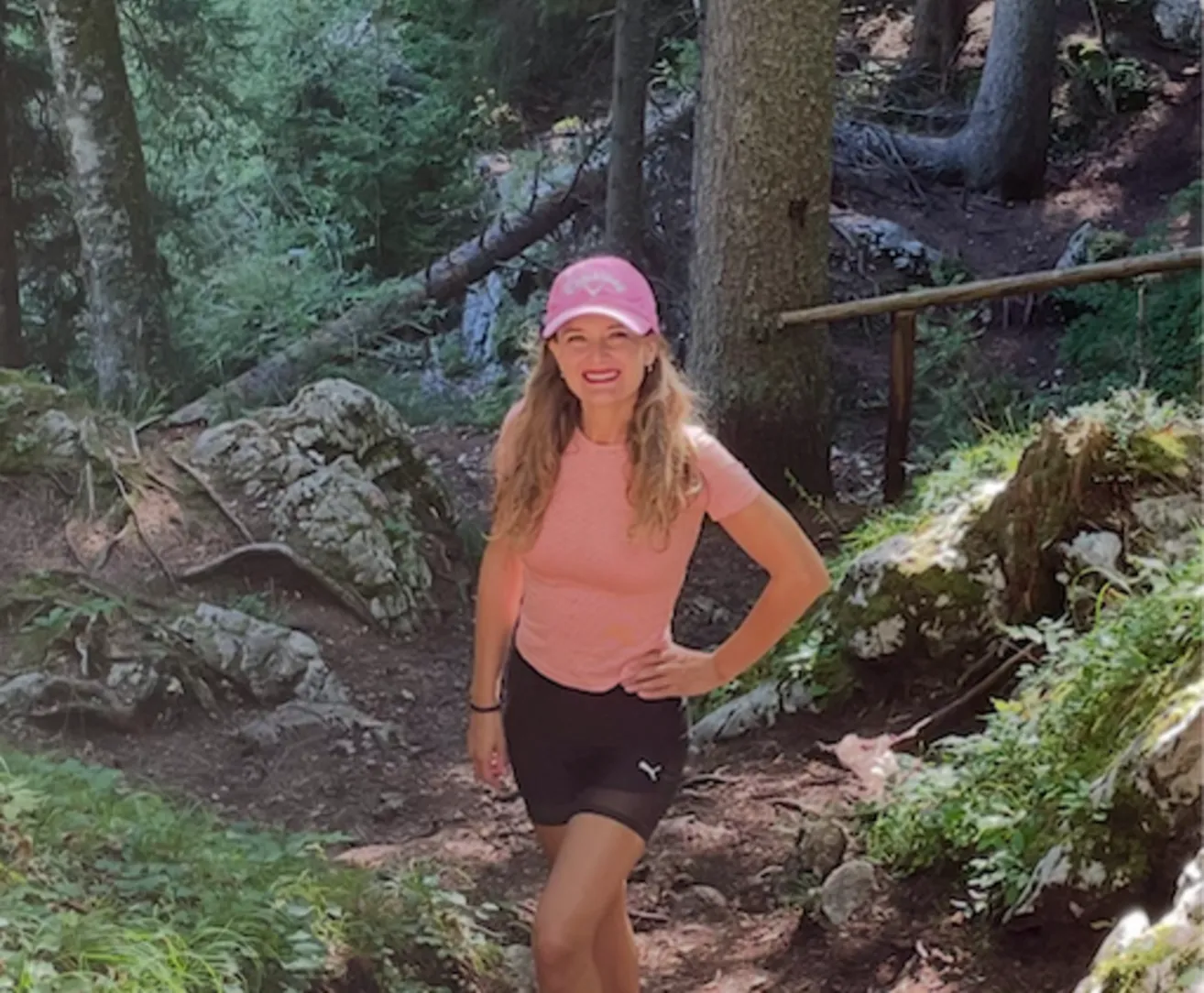 Eine Frau in einem rosa Hut und Shirt steht auf einem Wanderweg, lächelt und posiert für ein Foto.