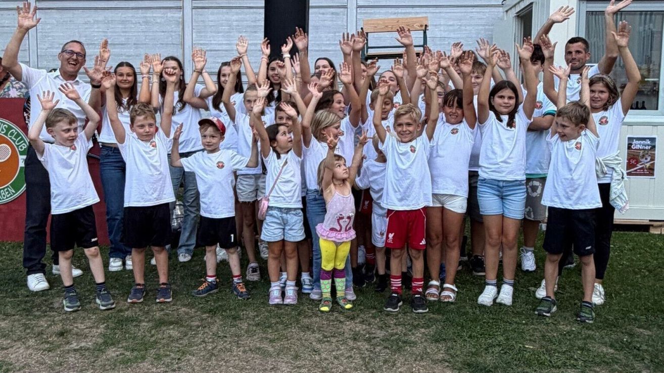 Eine Gruppe von Kindern in gleichen weißen T-Shirts hebt die Hände in die Luft, lächelt und posiert für ein Foto.