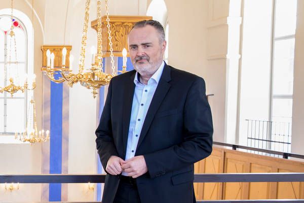 Ein Mann im Anzug steht in einem großen Saal mit Kronleuchtern und einer blauen Säule.