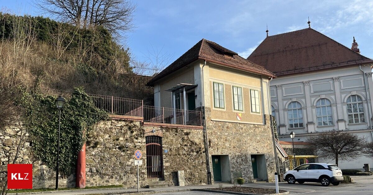 Ein kleines Steingebäude mit einem braunen Dach und mehreren Fenstern, umgeben von einer Steinmauer mit einem Metallzaun. Ein Schild ist in der Nähe des Eingangs an der Wand befestigt.