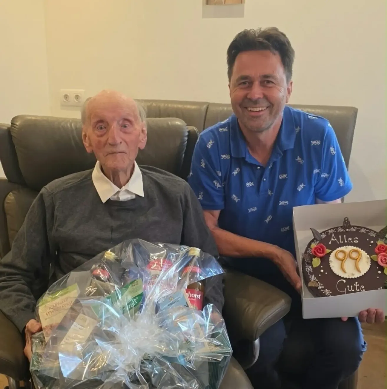 Ein älterer Mann und ein jüngerer Mann sitzen zusammen, der jüngere Mann hält einen Geburtstagskuchen mit der Nummer 99. Der ältere Mann hält eine Geschenktüte.