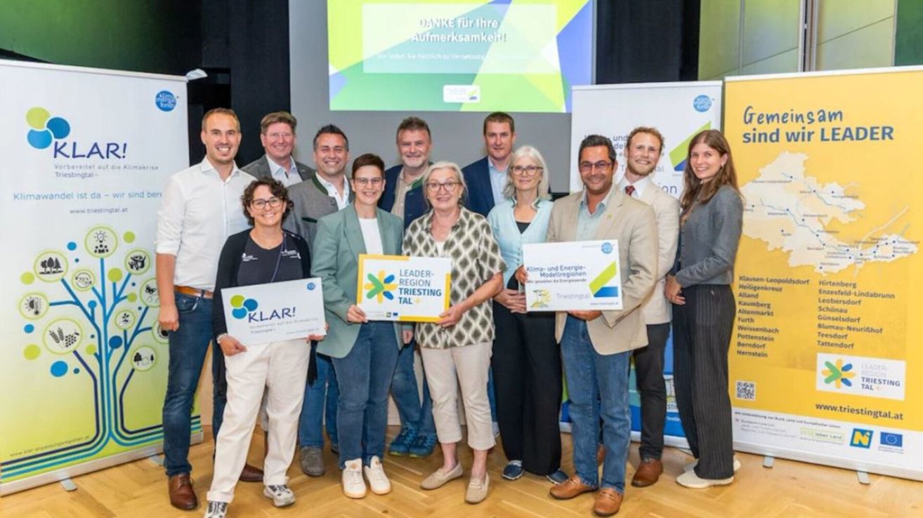 Eine Gruppe von Menschen steht zusammen und hält Schilder mit den Aufschriften 'KLAR!' und 'Leader-Region Triesting Tal.' Sie lächeln und scheinen bei einer Veranstaltung zu sein. Hinter ihnen befindet sich eine Projektionsleinwand mit einer Botschaft.