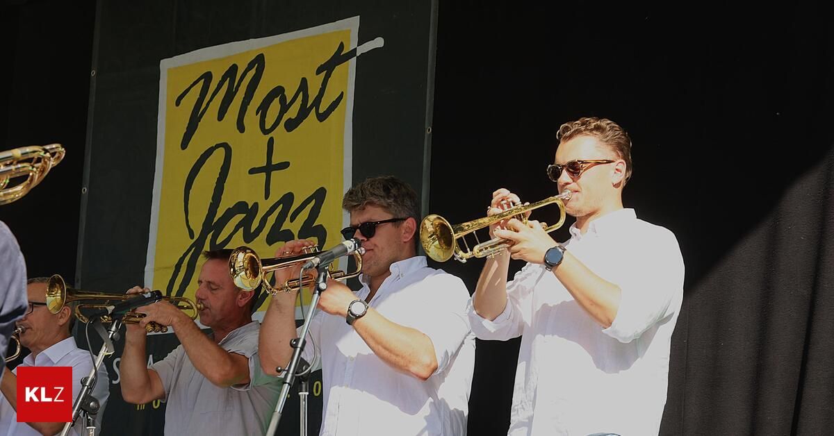 Vier Männer spielen Trompeten auf einer Bühne mit einem gelben Banner, das 'Most Jazz' steht. Die Männer tragen weiße Hemden und Sonnenbrillen. Der Mann auf der rechten Seite trägt eine Uhr.