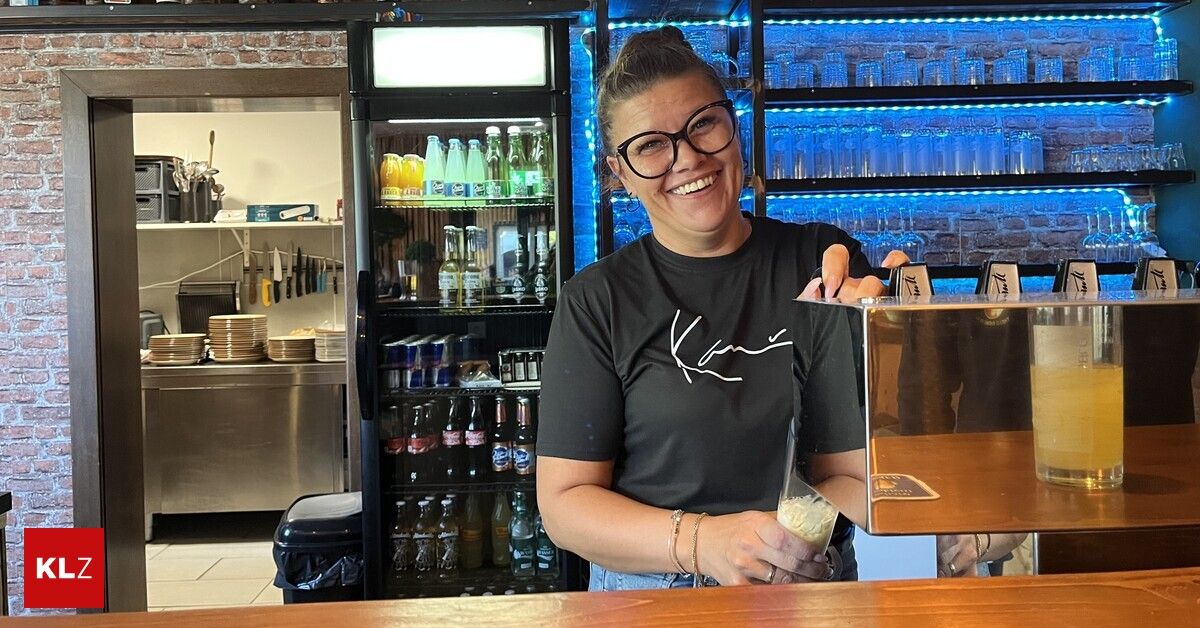Eine Frau mit Brille und einem schwarzen T-Shirt mit weißem Logo lächelt und hält ein Glas in der Hand in einer Bar.
