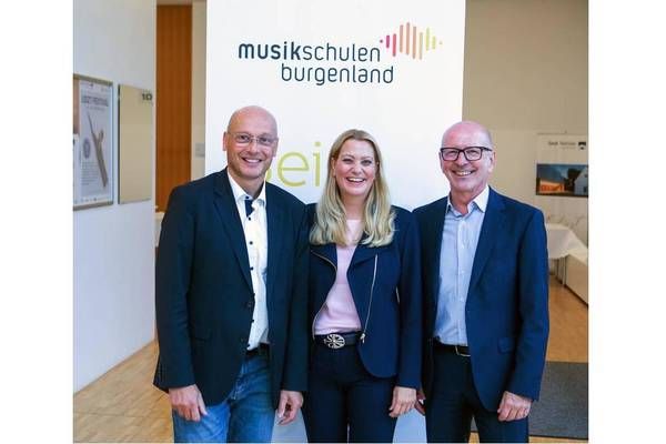 Drei Personen, zwei Männer und eine Frau, stehen zusammen vor einem weißen Banner mit dem Text 'musikschulen burgenland'. Sie alle lächeln und scheinen für ein Foto zu posieren.