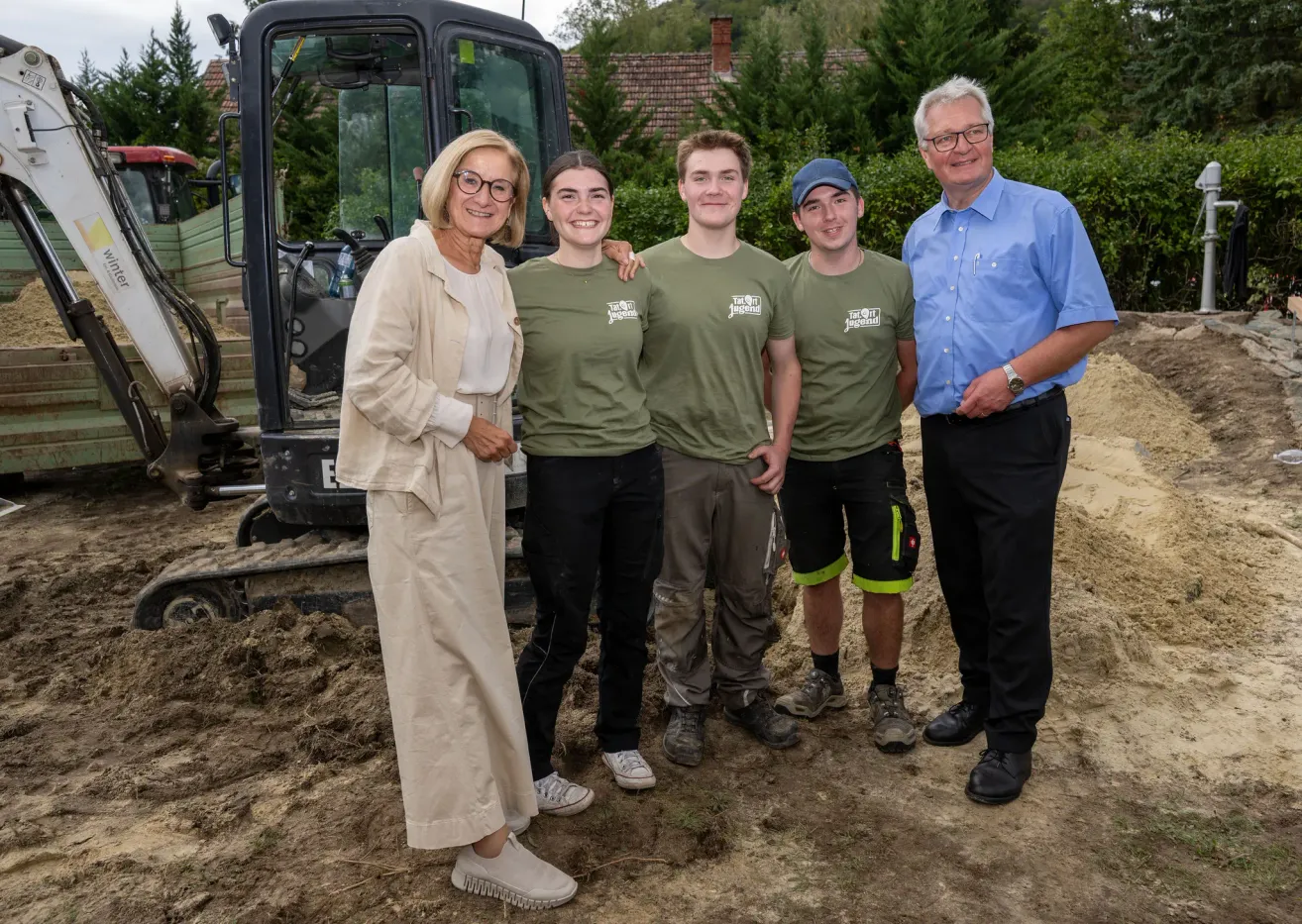 Eine Gruppe von Menschen in grünen T-Shirts und Turnschuhen steht auf einem schlammigen Gelände mit einem Baufahrzeug im Hintergrund.