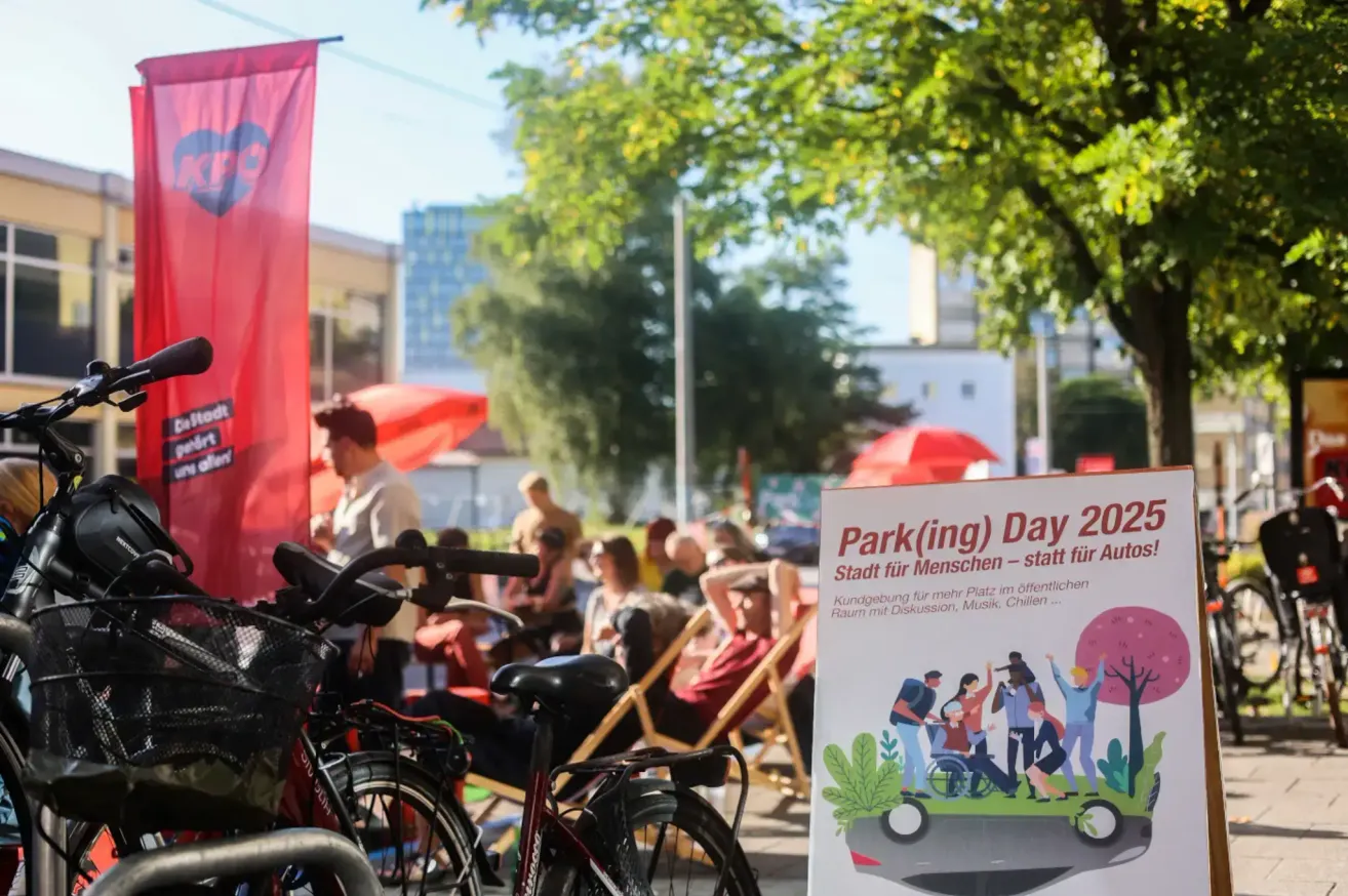 Ein lebhaftes Outdoor-Event mit Fahrrädern, Menschen und einem Schild für den Park(ing) Tag 2023. Das Schild wirbt für eine Stadt für Menschen, mit Diskussionen, Musik und Kindern.