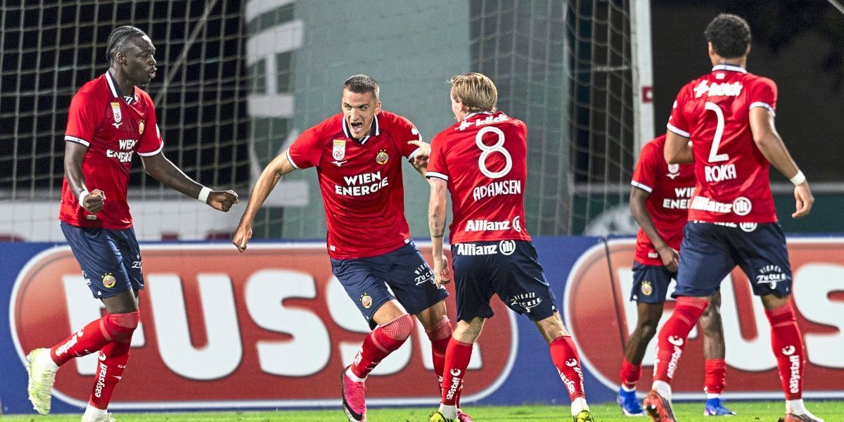 Zwei Fußballspieler feiern ein Tor auf einem Feld. Einer trägt ein rotes Trikot mit der Nummer 8, der andere ein rotes Trikot mit dem Text 'WIEN ENERGIE'. Sie sind von anderen Spielern umgeben.