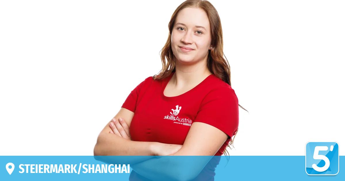 Eine Frau mit braunen Haaren, die ein rotes T-Shirt mit dem Text 'skillsAustria' und einem weißen Logo trägt. Sie lächelt und posiert für ein Foto mit verschränkten Armen.