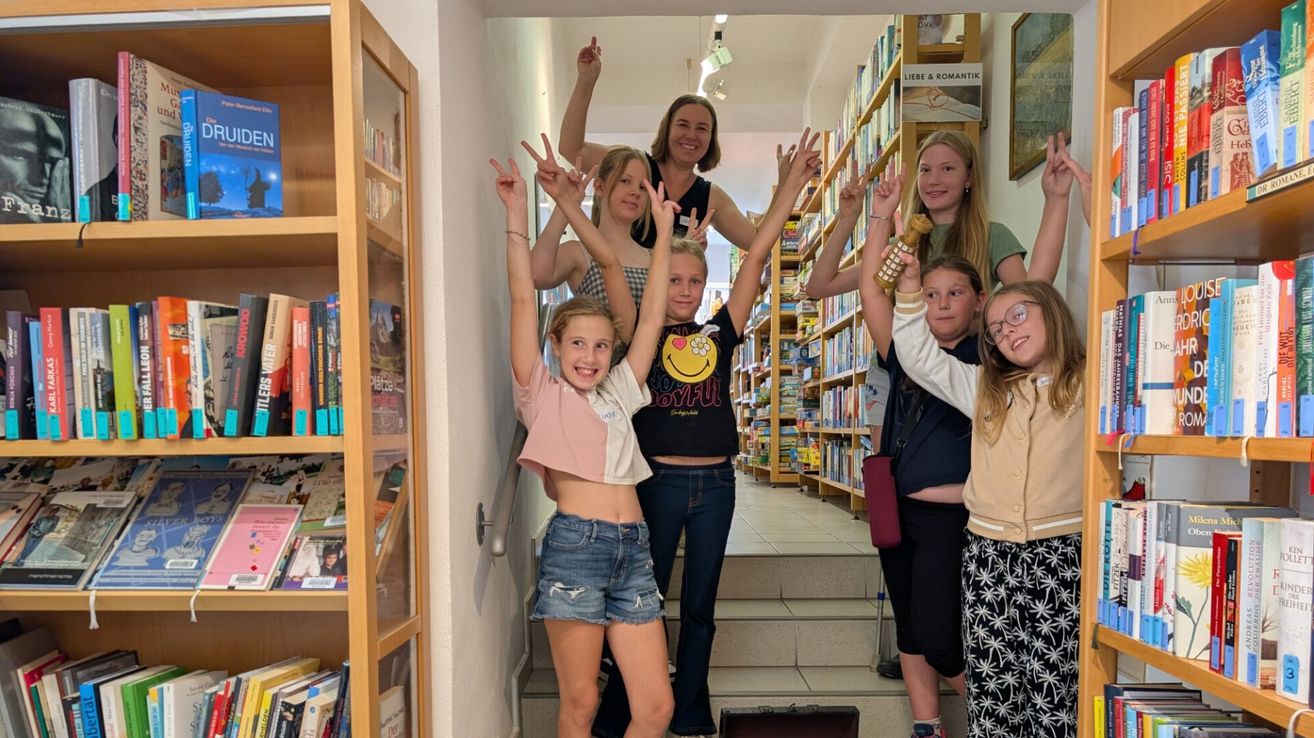 A group of young girls and women pose for a photo in a bookstore, with some making peace signs and others smiling.
