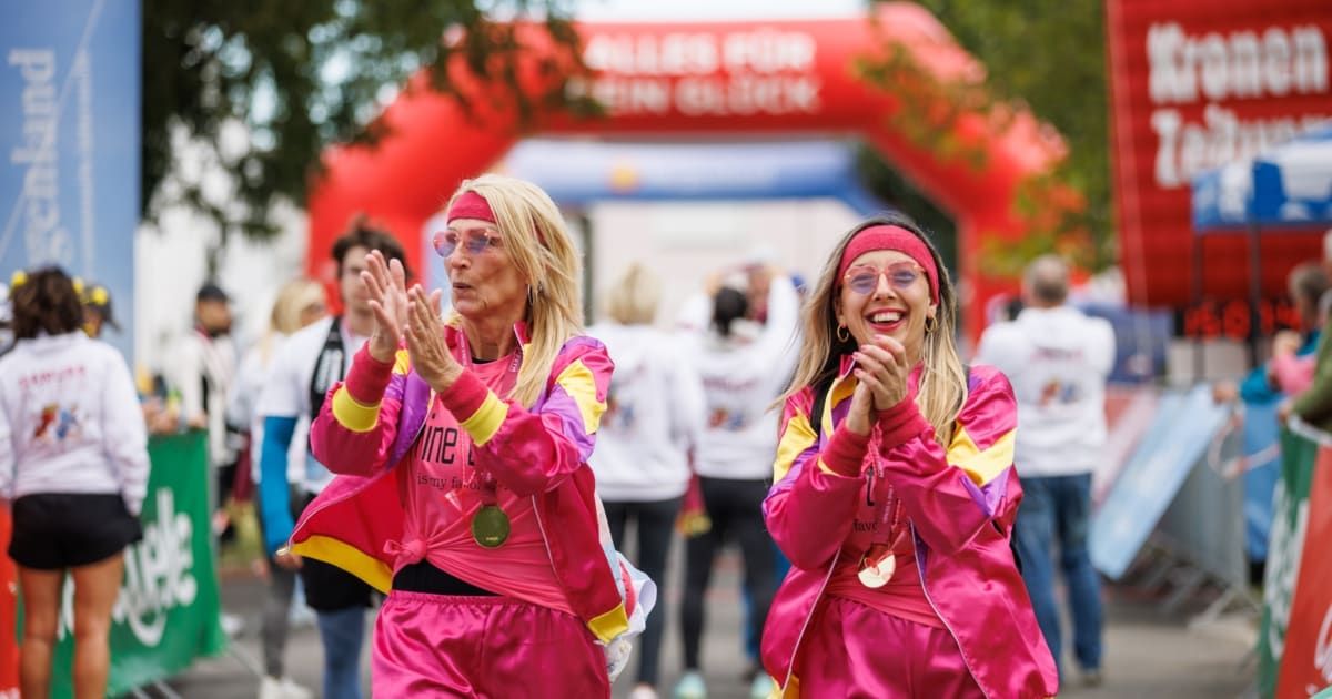 Two women in pink and yellow tracksuits celebrate at a marathon finish line.