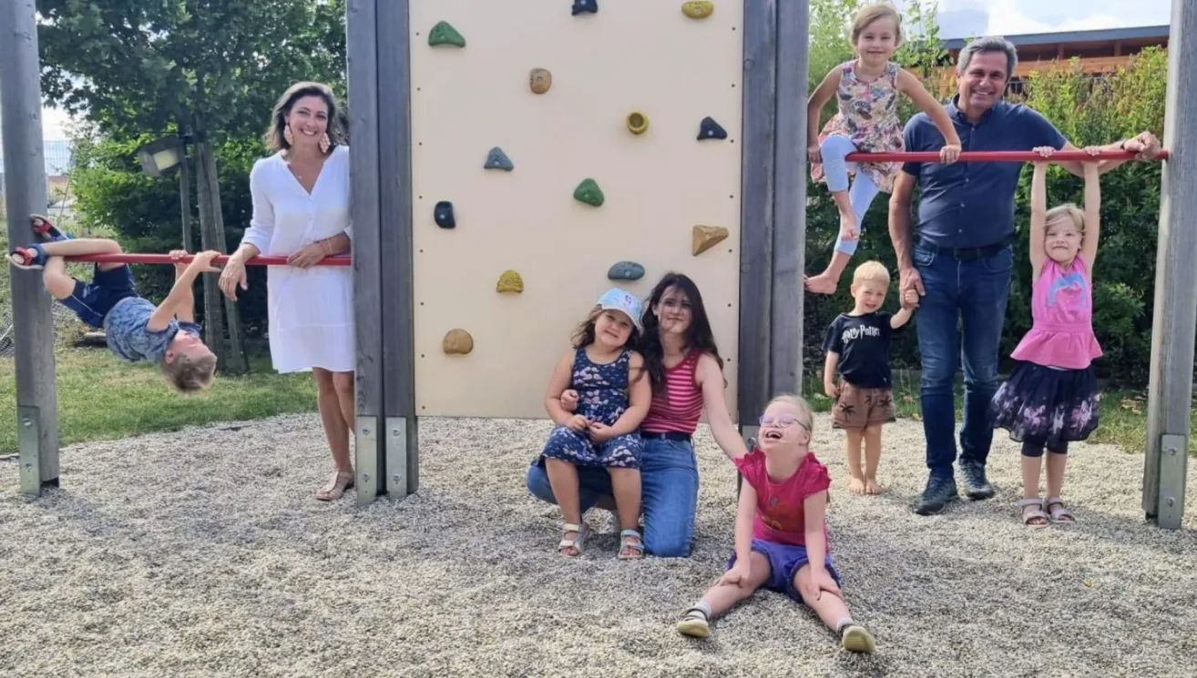 Eine Familie versammelt sich um eine Kletterwand in einem Park, eine Mädchen klettert und andere beobachten.