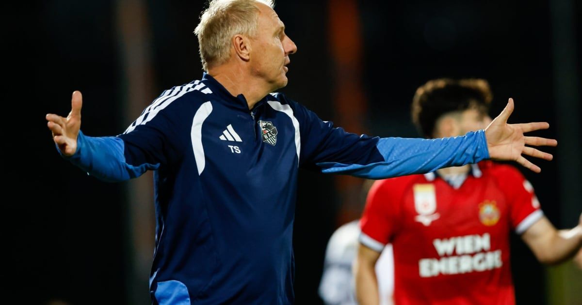 Ein Fußballtrainer in einem blauen Adidas-Jacke gestikuliert während eines Spiels mit den Armen.