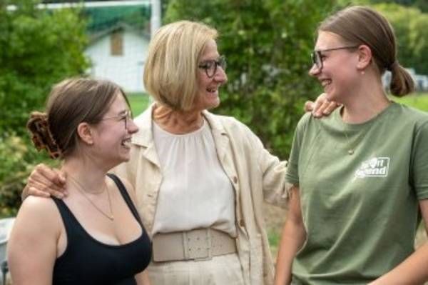 Drei Frauen, eine ältere mit Brille und zwei jüngere, eine in einem grünen Shirt, stehen zusammen und lächeln in einem Garten.